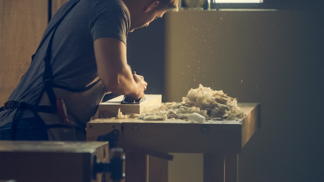 A craftsman skillfully shaping wood at a workshop bench with wood shavings visible.